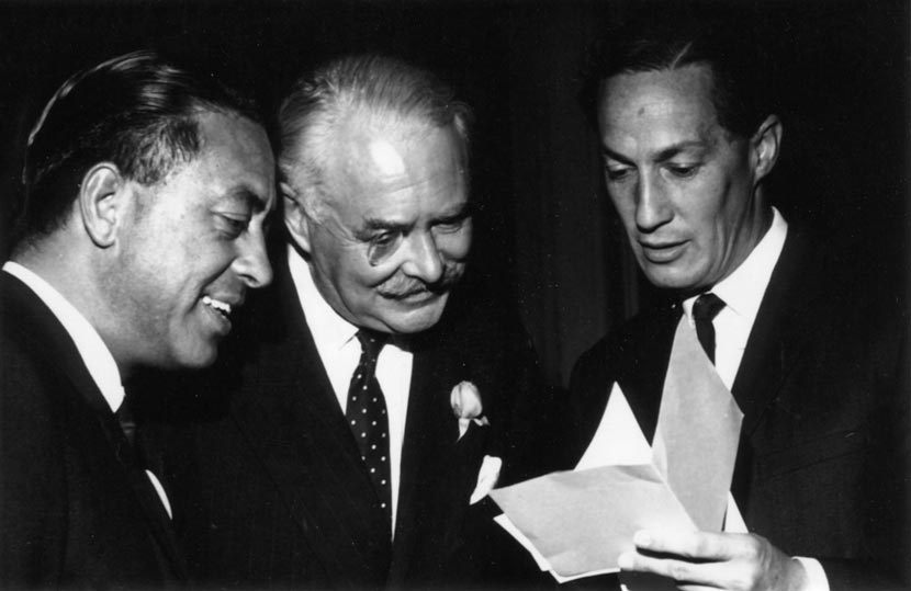 Three men look at a sheet of paper one of them is holding up. Fergusson, in the centre, wears a monocle, flanked by two Māori men in suits. The man on the left is unidentified.