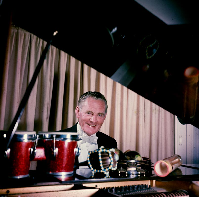 Elderly man in tuxedo sitting behind piano with musical instruments in the foreground.