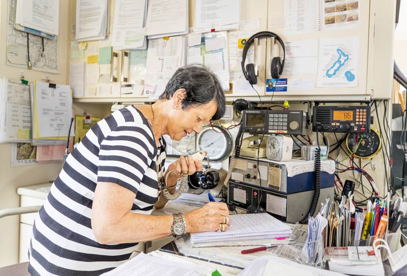 A woman talking into a radio, surrounded by electronic equipment and paper notes hanging on the wall.