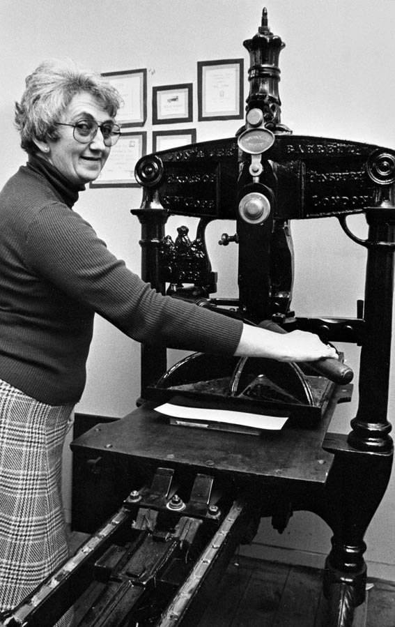 Mina McKenzie standing beside an antique printing press, turning a handle for the camera.
