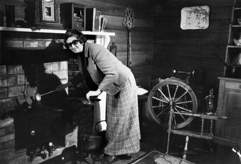 Mina McKenzie stands in a museum exhibit portraying the interior of a colonial house, with a fireplace, spinning wheel and other furniture. She pretends to ladle food from a pot for the camera.