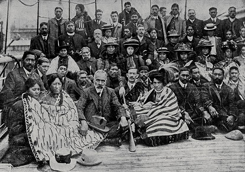 A group of Māori, dressed in European clothing, are seated on the deck of a ship. A woman in the front row holds a tewhatewha (long handled wooden weapon).