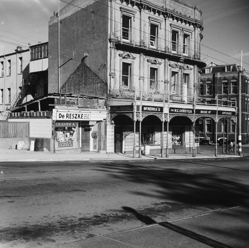 A two-storey brick building with Victorian architectural features, labelled McKenna’s House of Antiques, alongside a small, semi-derelict shop selling cigarettes. 