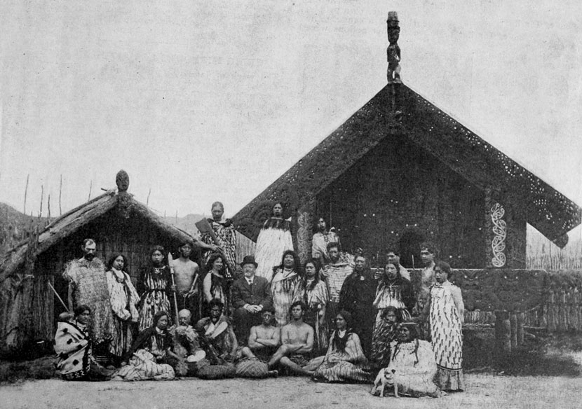 A group of Māori, dressed in traditional clothing, stand and sit outside a raupō whare and carved building, with a man wearing a European suit at the centre.