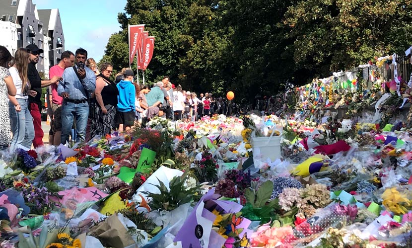 Line of people looking at ground covered in coloured flowers, handwritten cards and messages in front of a metal spiked fence