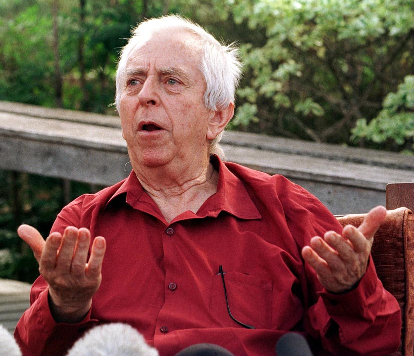 A man seated outdoors, gesticulating with his hands.