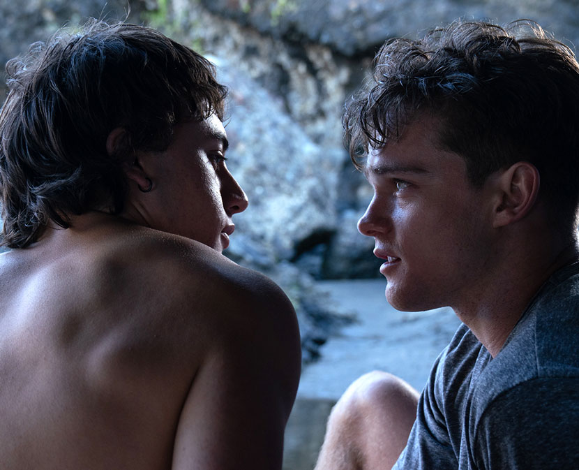 Two young men, one shirtless, staring into each other's eyes intimately.
