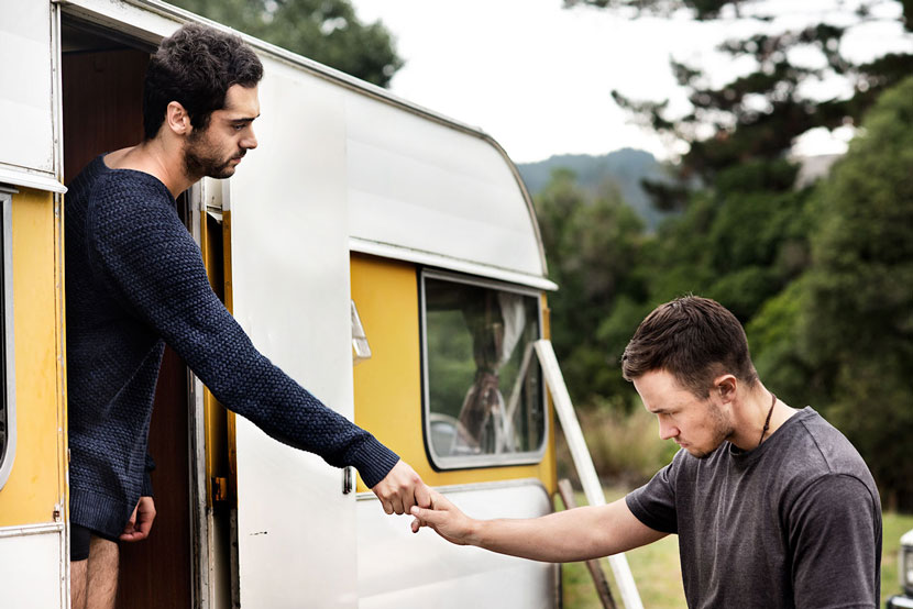 Man standing in door of caravan holding hands with another man with his head bowed