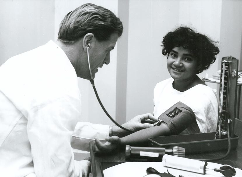 A doctor in a white coat conducts a blood pressure test on a young child, who smiles at the camera.