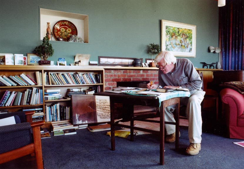 A man seated writing at a desk, alongside shelves of books, with several paintings visible.