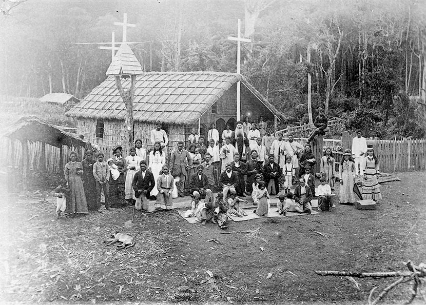 James McDonald and a large group of Māori in front of a thatched church building in a Māori village.