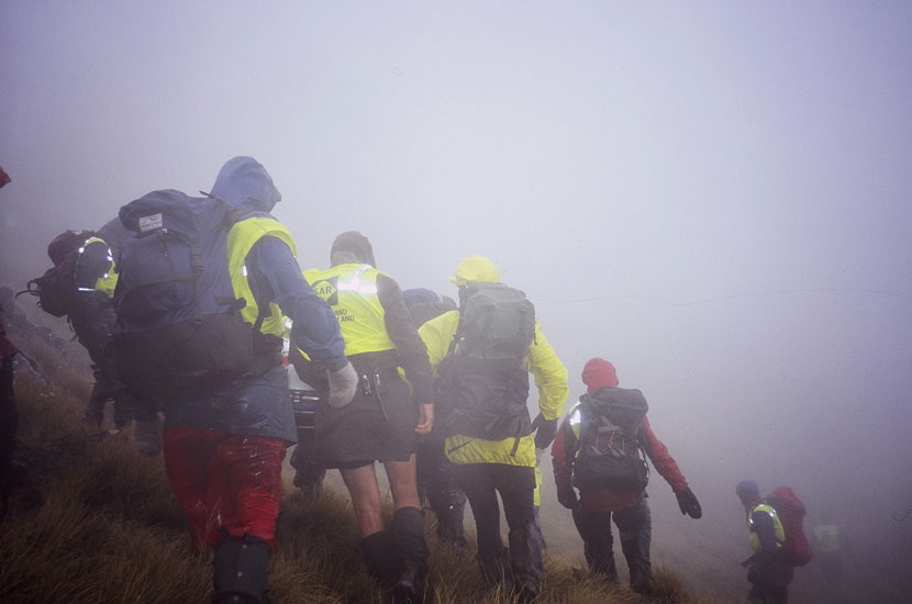 A large group of people in tramping clothing walk on a steep hill in fog. 