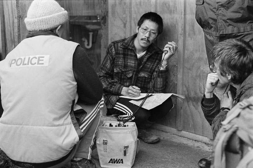 Men crouching around a radio, one listening to a radio transmitter. 
