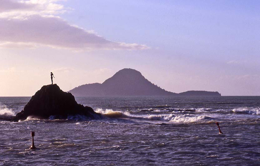 Statue of young girl atop a rock surrouned by sea with an island in the background.