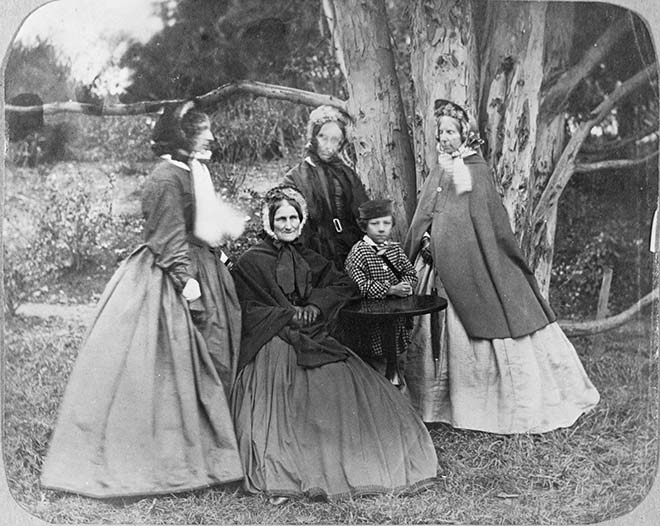 A group of formally-dressed women and a boy under a tree, some seated and some standing.