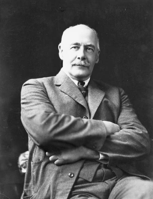 Kenneth Williams; bald, moustachioed, wearing a suit and tie, seated with arms crossed.