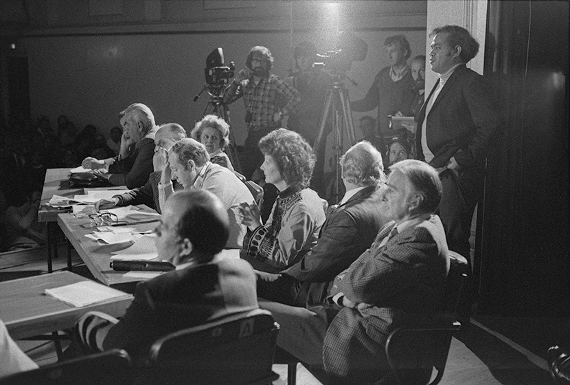 People sitting around meeting room with speaker standing and cameras in background