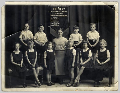Banner for 'H.M.C St John's Choir' behind group of children seated each side of a woman