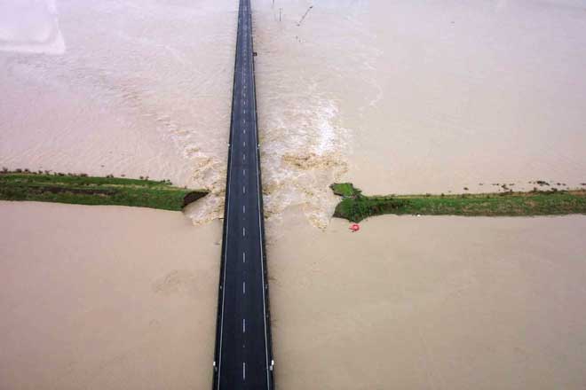 A road runs through flood waters with a broken stopbank on either side. 