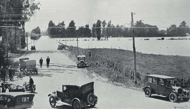 Cars and people collect together near a flooded river.
