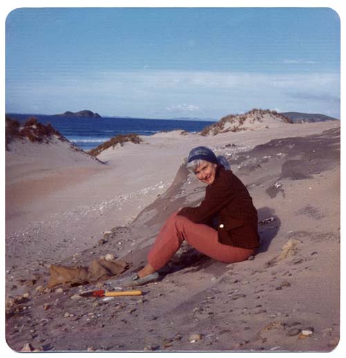 Woman sitting on beach. A trowel and sack are by her side