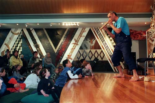 Barefooted man playing a wooden flute on a stage watched by a crowd of children and adults.