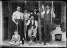 Group of men on porch holding up two enormous eels. A boy sits in front