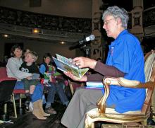 Woman seated on stage reading to audience parents and children