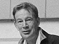 A photograph of Ian Athfield seated at the bottom of a paved ramp with a roof supported by columns sculpted to look like nikau palms